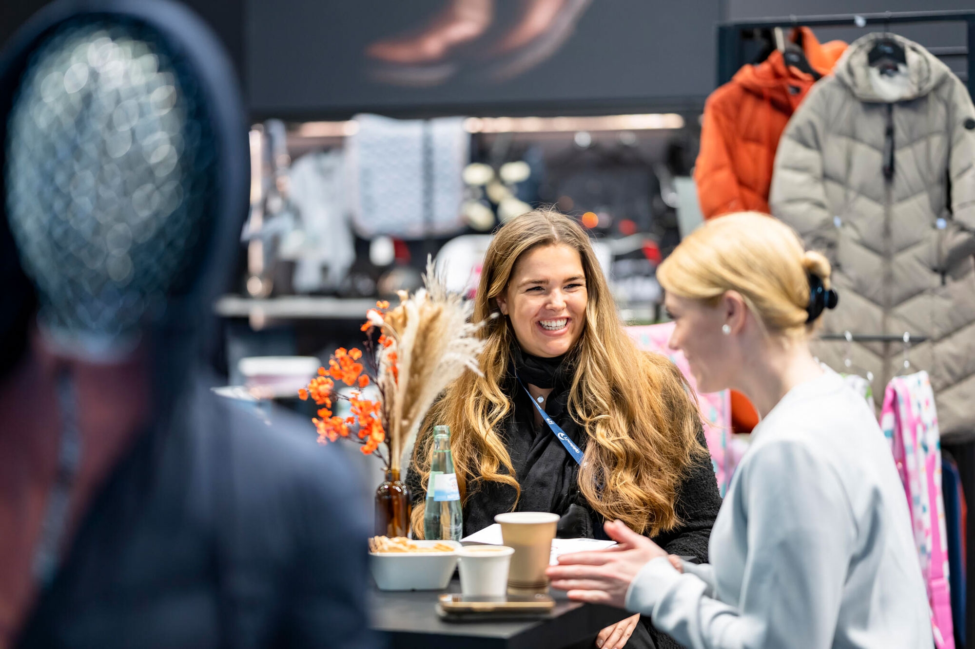 Event Area Two women sit at a table with coffee cups and a glass bottle, jackets hanging on a coat rack in the background.