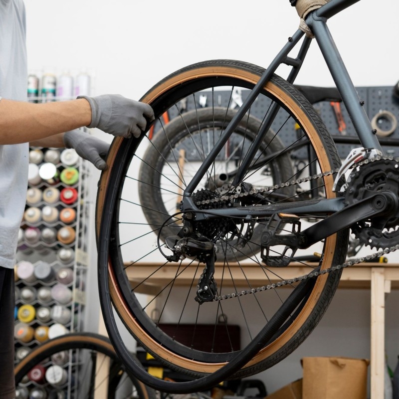 Eine Person mit Handschuhen repariert das Rad eines Fahrrads in einer Werkstatt, umgeben von Fahrradteilen und Werkzeugen, die ordentlich auf Regalen angeordnet sind.