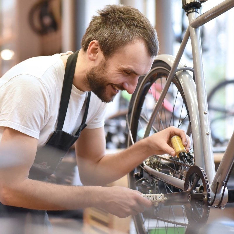 Ein Mechaniker lächelt, während er in einem gut beleuchteten Fahrradgeschäft einen silbernen Fahrrad repariert, Werkzeuge benutzt und Komponenten anpasst, um erstklassige Dienstleistungen anzubieten.