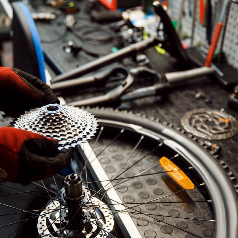 Eine Person in roten Handschuhen hebt ein Fahrradcassette über ein Rad, umgeben von Werkzeugen und Fahrradteilen auf einer Werkbank, die voller Ersatzteile ist.