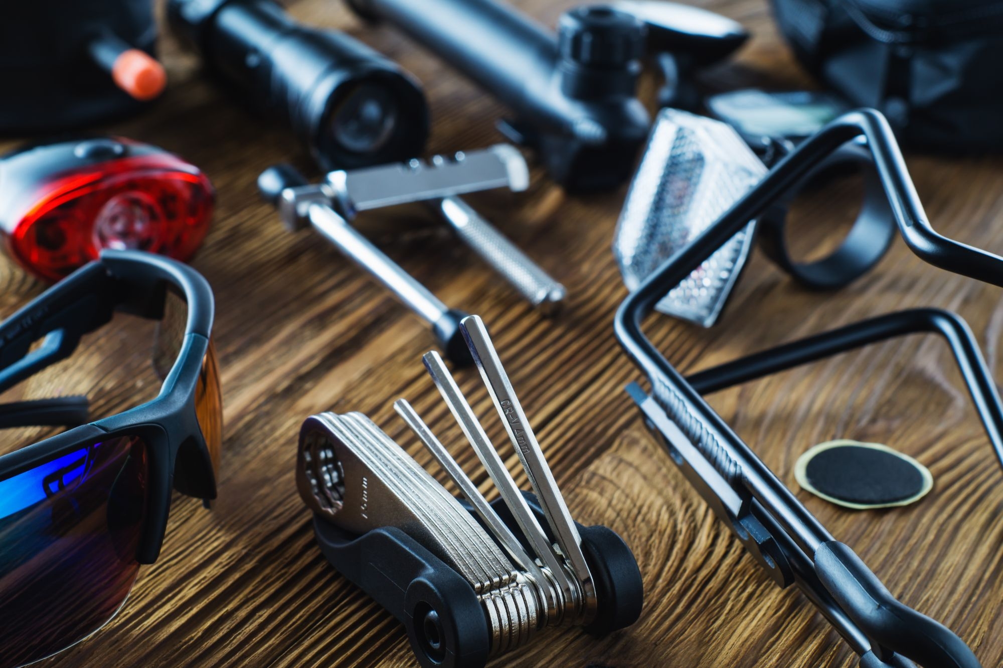 Eine Ansammlung von Radfahrwerkzeugen, Sonnenbrillen und Equipment, darunter ein Multitool, eine Taschenlampe und Fahrradzubehör, ist sorgfältig auf einer rustikalen Holzoberfläche angeordnet.